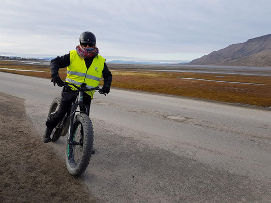 Een persoon in een geel reflecterend vest rijdt op een fatbike langs een onverharde weg in een bergachtig landschap met gras en water op de achtergrond.