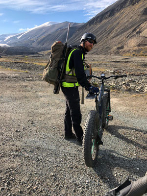 Een persoon met een rugzak staat naast een fatbike op een ruig terrein met bergen op de achtergrond.