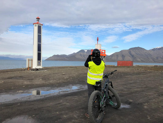 Een persoon met een veiligheidsvest staat op een fatbike, kijkend naar een markering nabij het water en bergen op de achtergrond.