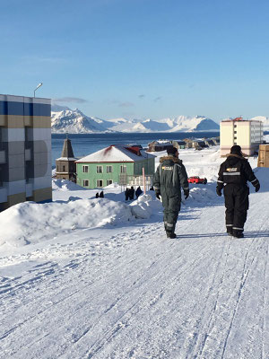 Twee mensen wandelen op een besneeuwde straat in Barentsburg, met kleurrijke gebouwen en bergen op de achtergrond.