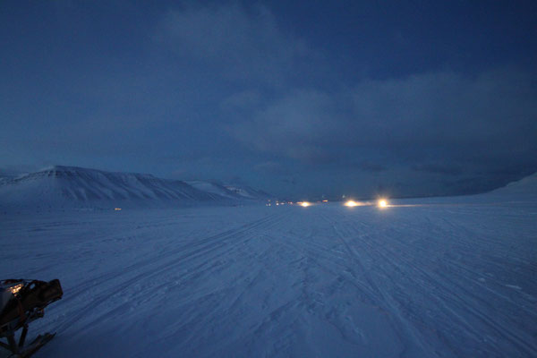 Een sneeuwlandschap bij nacht, met lichten in de verte en bergen op de achtergrond.