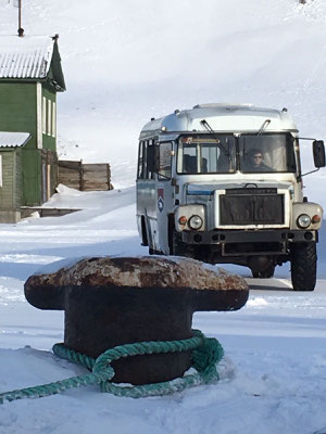 Een sneeuwbedekte omgeving met een oud voertuig in de verte, en een verroest anker met touwen op de voorgrond, nabij een gebouw in Barentsburg.