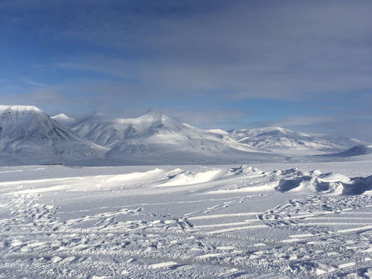 Een uitgestrekt sneeuwlandschap met bergen op de achtergrond en een blauwe lucht.