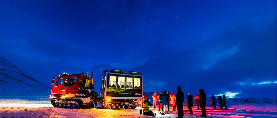 Een groep mensen staat in de sneeuw bij een sneeuwcat in een nachtelijk landschap met een blauwe lucht.