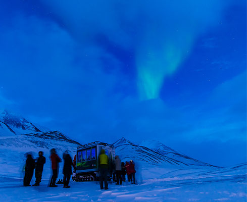 Een groep mensen bewondert het noorderlicht boven een besneeuwd landschap terwijl ze bij een sneeuwcat staan.
