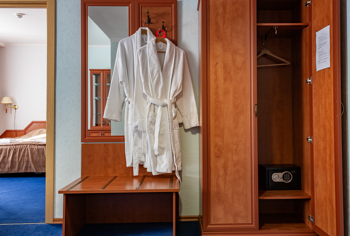 Interieur van een hotelkamer in Barentsburg met een spiegel, een houten kapstok en witte badjassen, een kast met een kluis en een bed op de achtergrond.