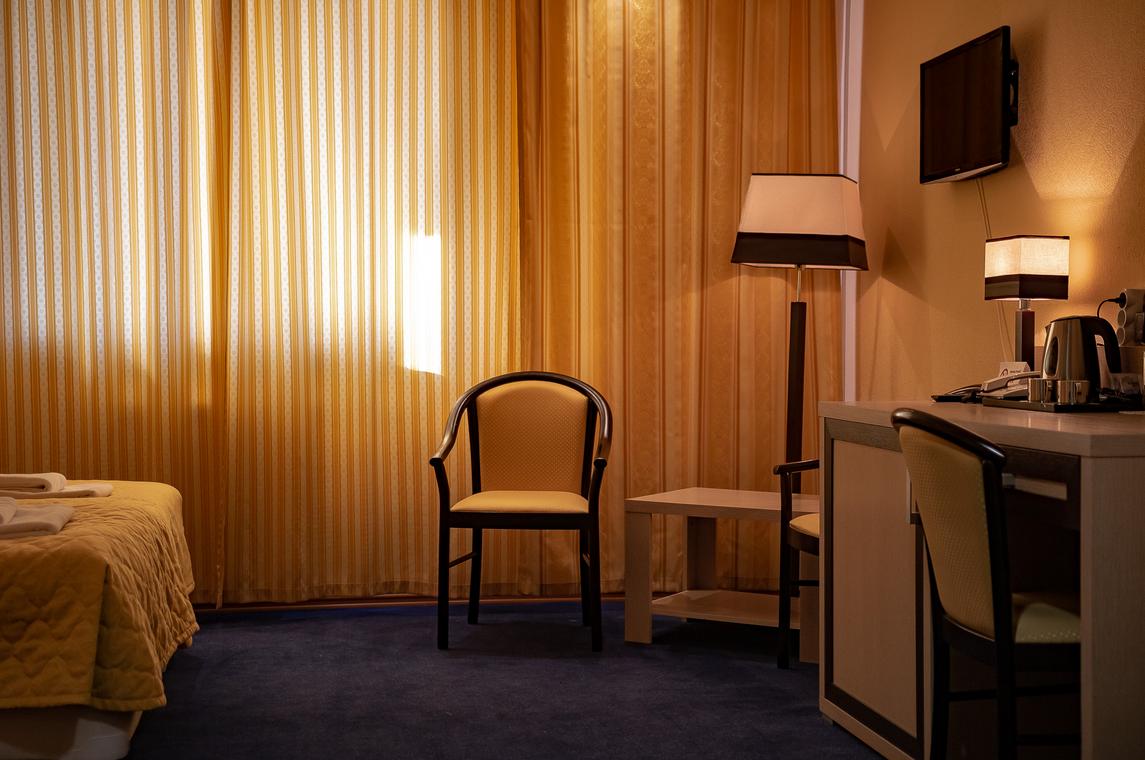 Een gastenkamer in Hotel Barentsburg met een bed, bureau, stoelen en grote raambekleding die het ochtendlicht filtert.