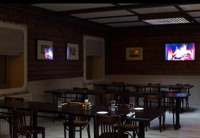 Een interieur van een restaurant in Barentsburg met houten muren en tafels, voorzien van glazen en borden. Op de muren hangen foto's en er zijn schermen met een afbeelding van een open haard.