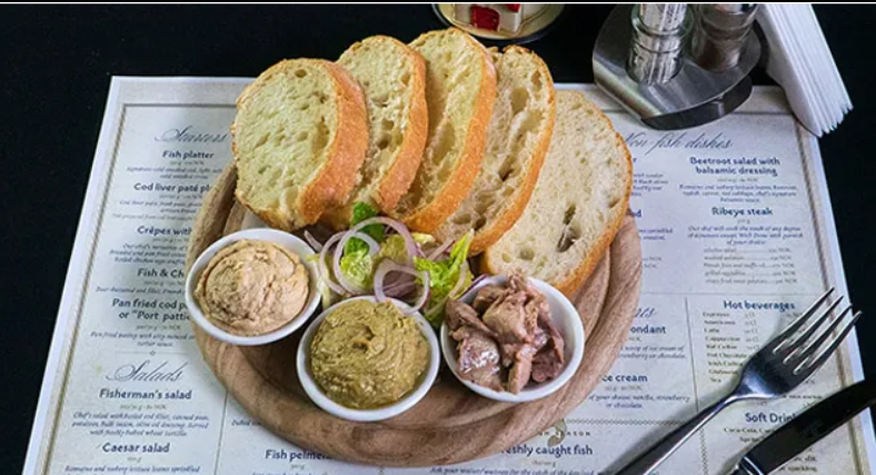 Een bord met versgebakken brood en drie kleine schaaltjes met visspread, geserveerd op een houten plank. Op de achtergrond is een menukaart zichtbaar met verschillende gerechten.
