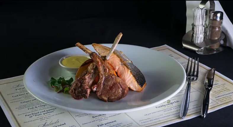 Een bord met een portie gegrilde zalm en lamskoteletten, geserveerd met een sausje en garnituur op een elegante manier, met restaurantmenu op de achtergrond.