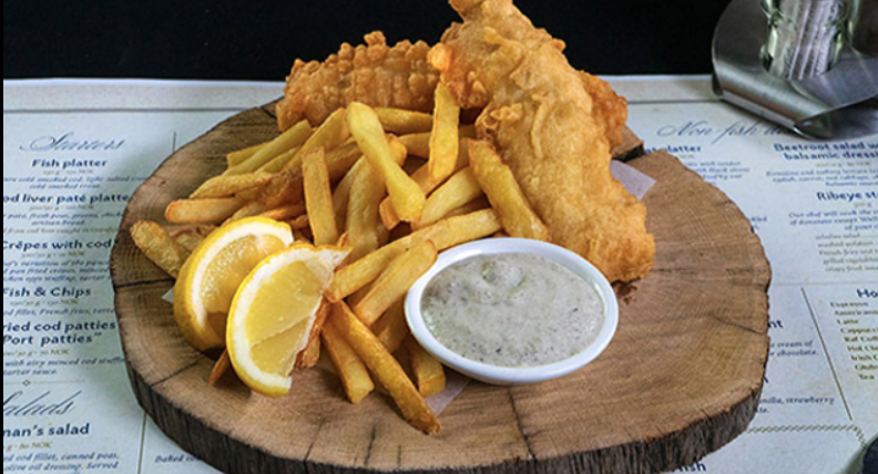 Een bord met fish and chips, bestaande uit krokante gefrituurde vis en goudbruine frietjes, geserveerd met schijfjes citroen en een kommetje saus op een houten plank.