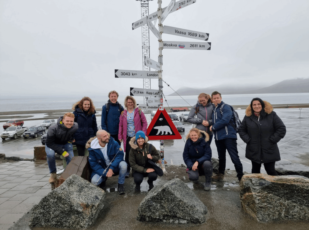 Groepsfoto van deelnemers aan een reis in Spitsbergen, voor een richtingbord met afstanden naar verschillende steden, waaronder een waarschuwingsbord voor ijsberen. Achtergrond toont een kustlijn met een zee onder een bewolkte lucht.