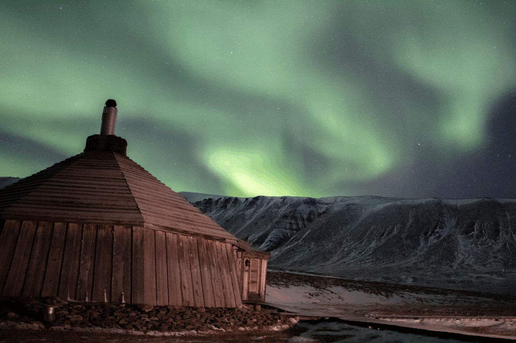 Noorderlicht boven een houten hut in Spitsbergen, met bergen op de achtergrond.