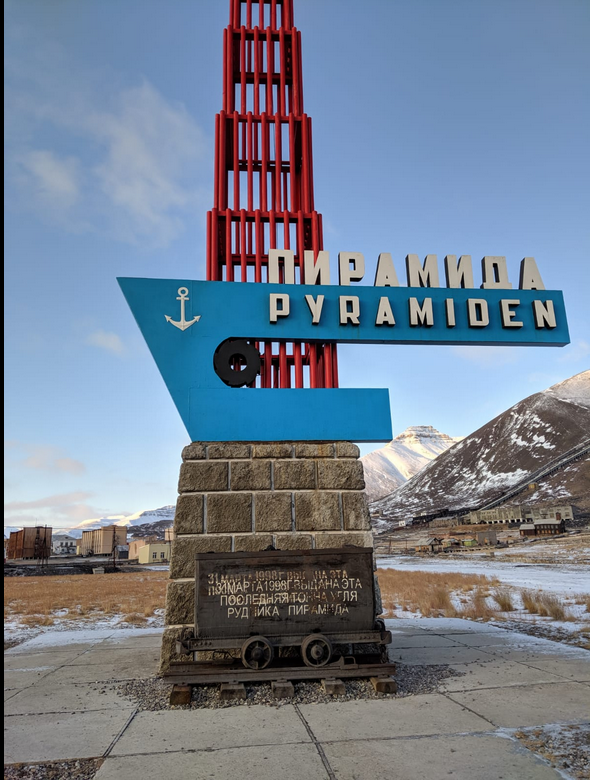 Een kleurrijk welkomstbord van Pyramiden, met de tekst 'PYRAMIDEN' in blauw en de Russische tekst erboven, omringd door bergachtige achtergrond en een rustige, open omgeving.
