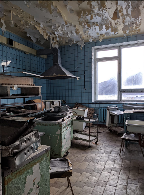 Een vervallen keuken met groene kasten en apparatuur, beschadigde muren en een uitzicht op de buitenomgeving.