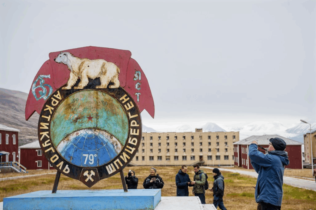 Een groep mensen staat bij een metalen sculptuur van een ijsbeer dat het noordpoolgebied symboliseert, met opvallende gebouwen en bergen op de achtergrond.