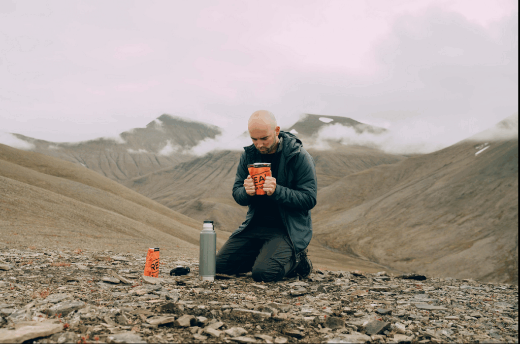 Een man knielt op een bergachtige ondergrond in Spitsbergen en houdt een warme drank vast, met uitlopend landschap en mistige bergen op de achtergrond.