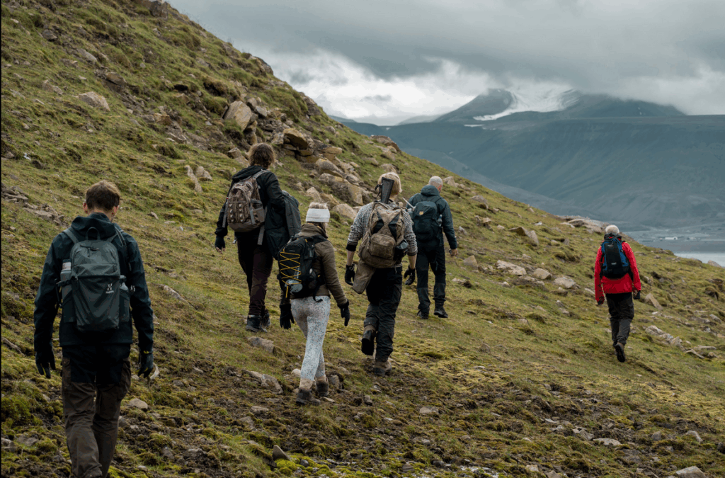 Een groep mensen aan het hiken op een groen heuvelachtig landschap in Spitsbergen, met bergen en een bewolkte lucht op de achtergrond.
