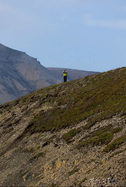 Een persoon met een camera staat op een glooiende heuvel in het Spitsebergen landschap, omgeven door bergen en groenige vegetatie onder een blauwe lucht.