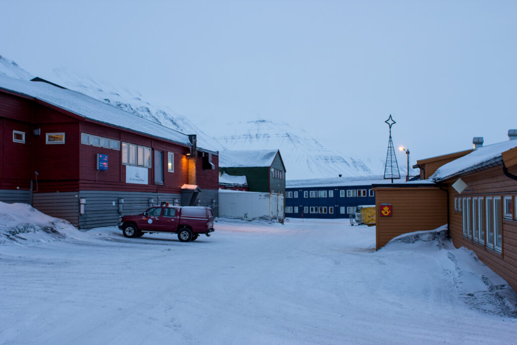 Sneeuwbedekte gebouwen en voertuigen in Longyearbyen, omringd door bergen onder een grijzige lucht.